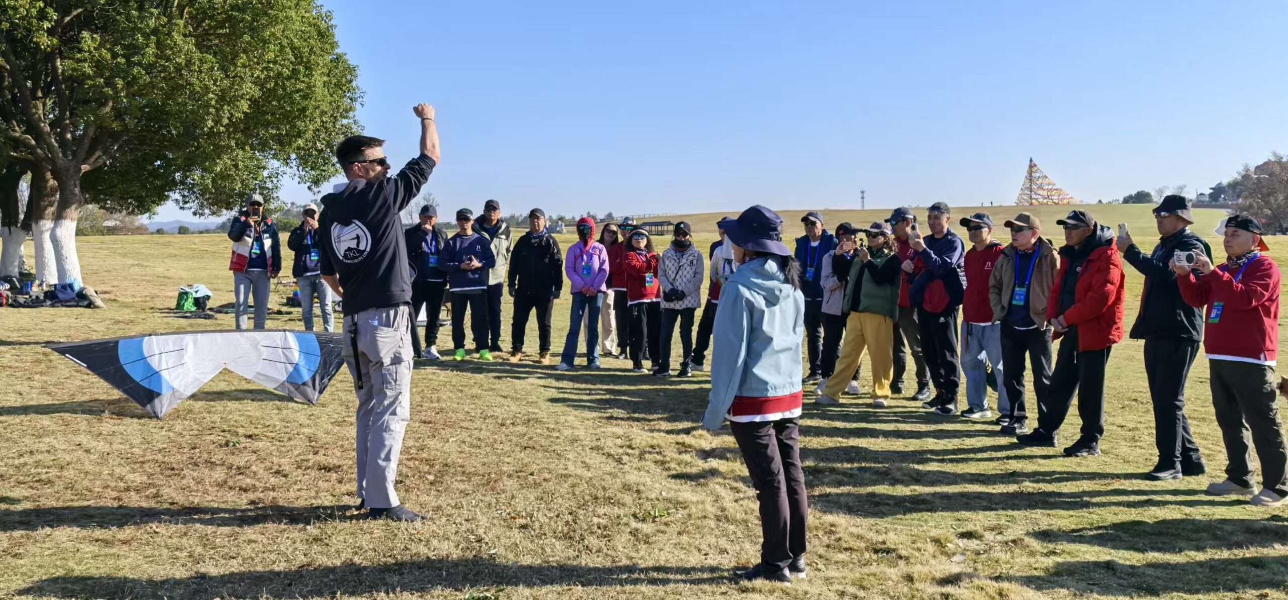 John Barresi addressing a large group of kite fliers on a grassy field, one arm raised, a dual line kite on the ground beside him