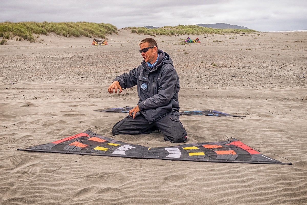 John Barresi kneeling on a beach beside two kites laid out on the sand